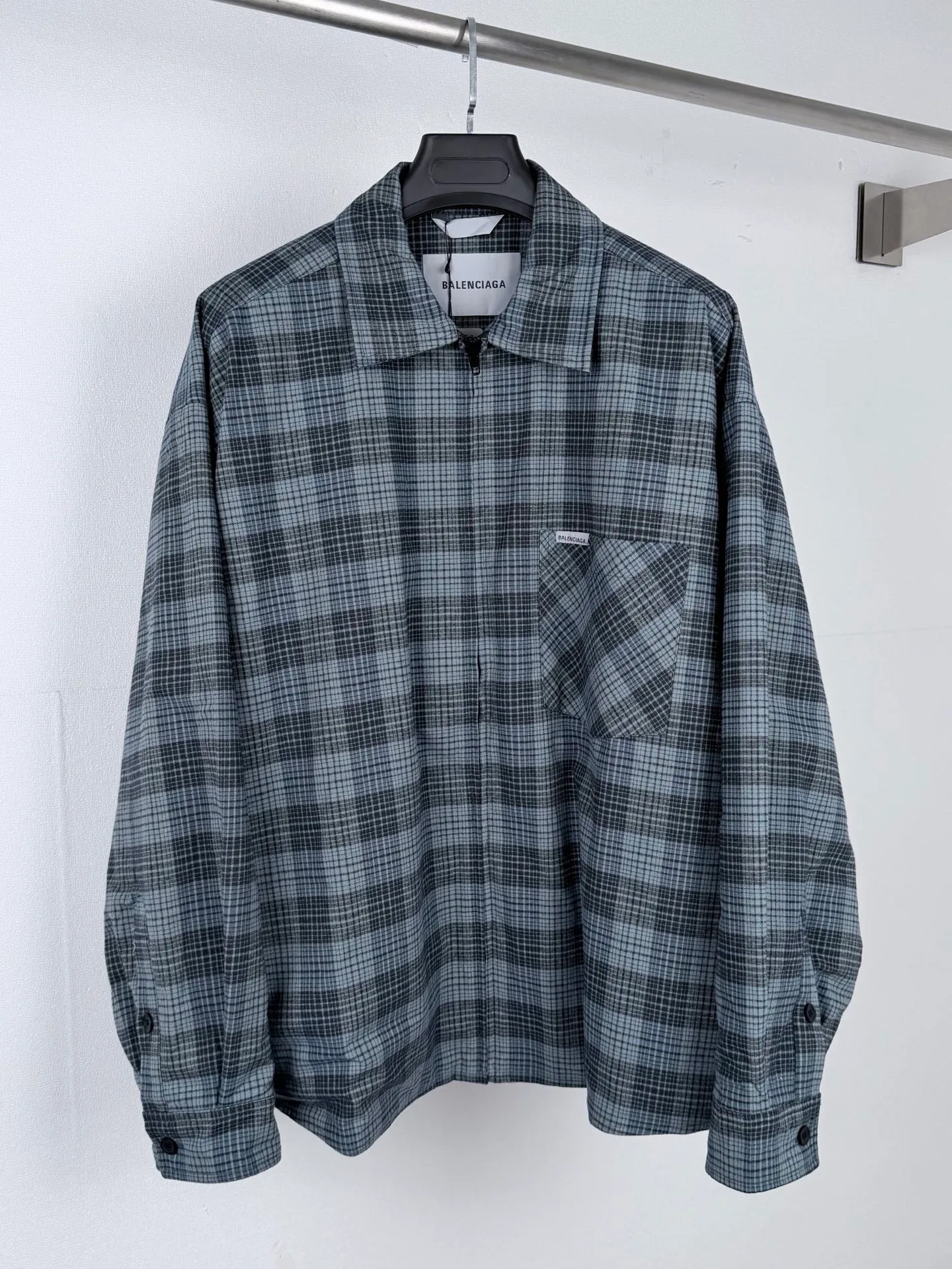 BALENCIAGA/balenciaga blue and gray plaid zipper pocket logo quilted jacket combines a three-layer structure of plaid woven fabric, filling cotton, and lining, and integrates plaid alignment technology and quilted garment production standards. There are multiple process difficulties in the whole process. The surface layer of the body is made of blue-gray Retro plaid fabric, and the cutting machine production strictly follows the global grid standards. The arrangement direction of the cutting pieces is fixed and cannot be changed at will. The horizontal and vertical lines of shoulder seams, side seams, and zipper placket joints must be consistent and coherent. The left chest patch pocket is cut independently, and the bag check pattern must be aligned with the front reference pattern. Slight deviations will destroy the visual integrity and directly increase fabric loss and cutting time. The most difficult part of the style is the three-layer composite sewing. The outer plaid fabric, filling cotton, and lining fabric have different physical properties, and the preshrinking processing parameters need to be adjusted separately. Pressure and temperature control are crucial during lamination operations. Once the parameters are out of balance, it can easily cause fabric stretch and distortion, plaid dislocation, and wrinkles and bulges in garments. At the same time, it is necessary to reasonably plan the quilting line to avoid the visual beauty of the grid pattern from being cut by the lines on the basis of firmly fixing the cotton layer and preventing cotton from running away. The placket adopts a centered zipper structure. It is difficult to sew the zipper tape and the quilted panels. It is required that the width of the left and right plackets be equal, the zipper routing is straight and smooth, and there is no lag in opening and closing. When the shirt lapel is filled with cotton, the thickness of the cotton needs to be controlled to prevent the collar from becoming bloated and unnaturally folded. The metal nameplate of the chest pocket is sewn at fixed points to avoid pulling the fabric during the operation and ensure that the plaid pattern is smooth and not deformed. The quilted material increases the overall thickness of the fabric. When sewing, you need to continuously adjust the presser foot pressure and stitch length to adapt to changes in sandwich thickness. The low-temperature and gentle ironing process is used for post-finishing to prevent the plaid from fading and the cotton layer from clumping due to high temperatures. From the precise alignment of cutting pieces, three-layer fabric pretreatment, composite quilting to garment finalization inspection, the processes are interlocking and the error tolerance rate is extremely low, testing the accuracy of plate making, equipment control and turning technology. Size:XS S M L